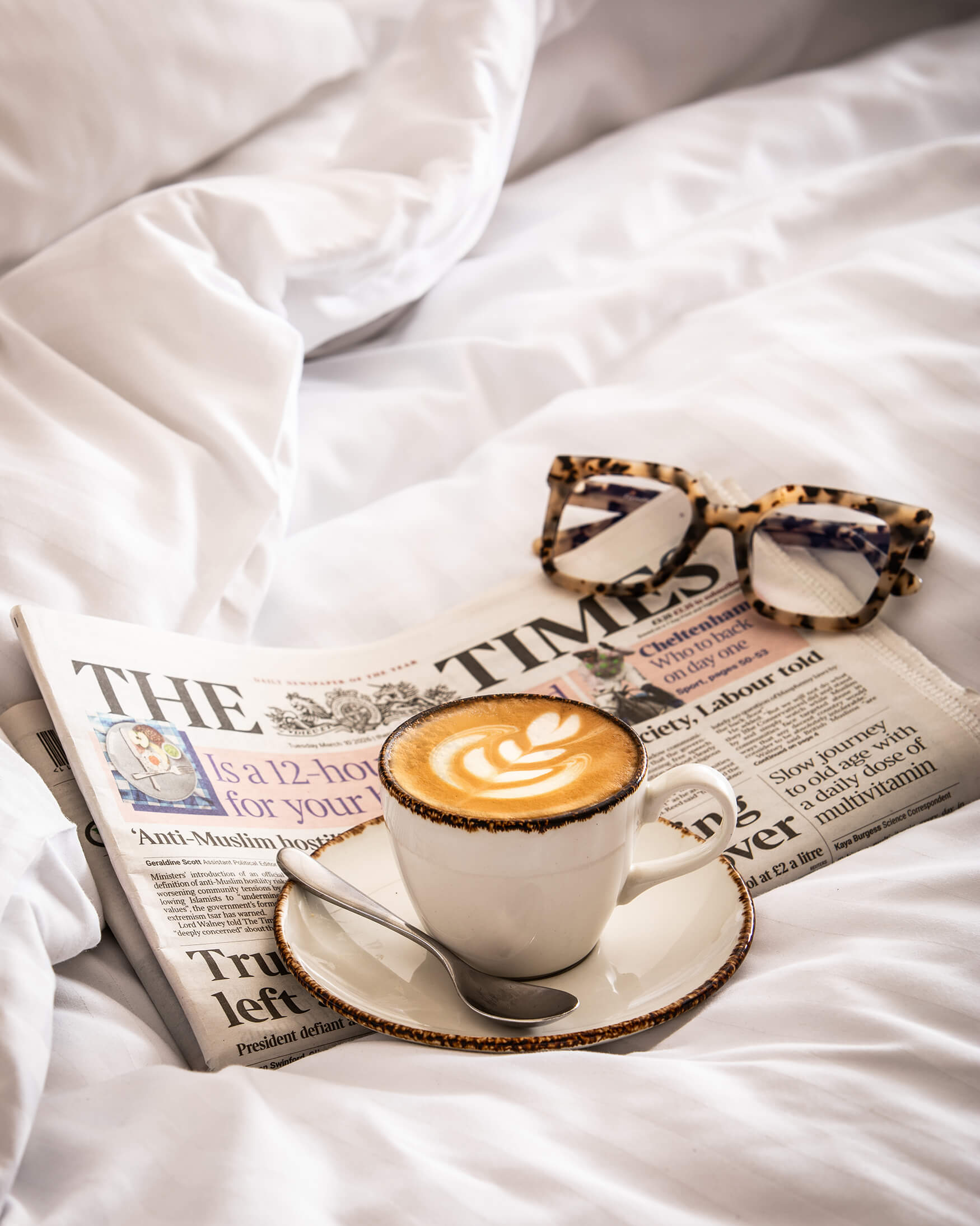 Image of a cup of coffee, The Times newspaper and some reading glasses on the crumpled bed of the rooms at The General Tarleton