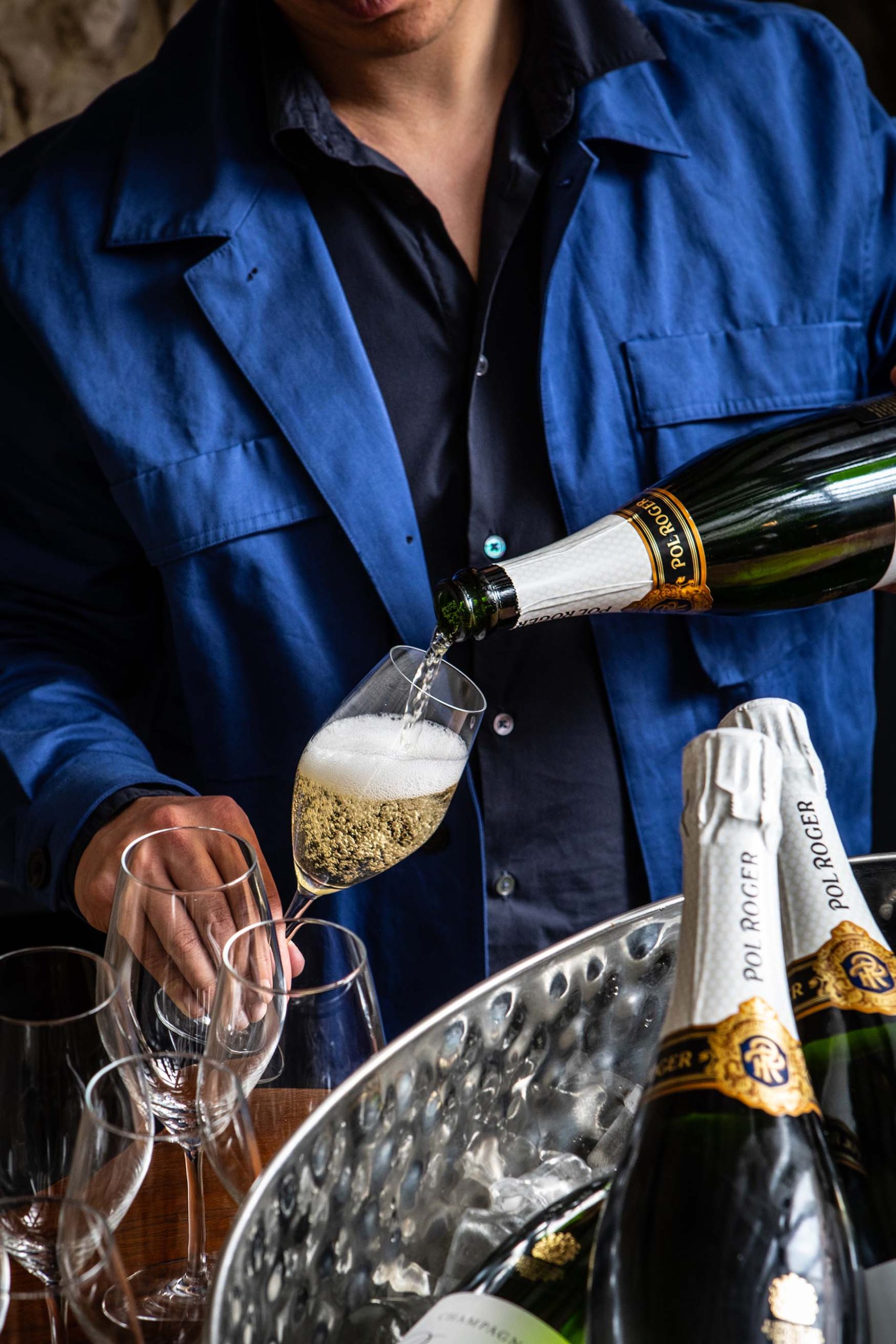 Image of a man in a blue workman's jacket pouring champagne into a glass