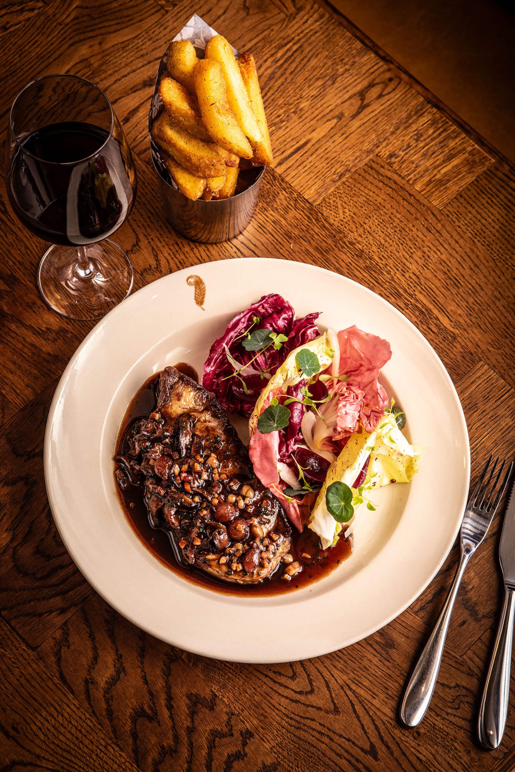 Overhead shot of a steak with salad and chips on a wooden table and a glass of red wine.