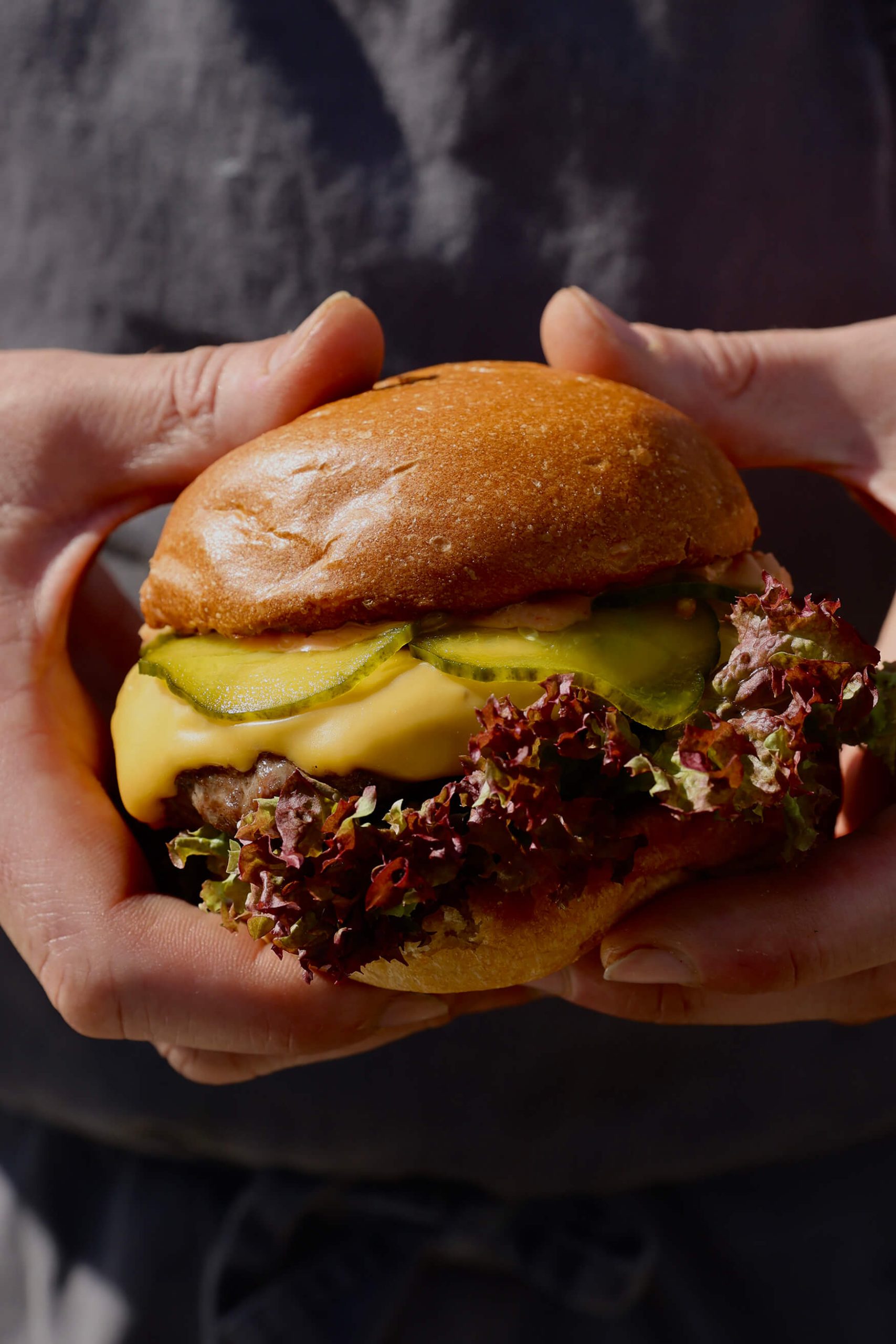Image of someone holding up a burger close to the camera showing oozing cheese, beautiful burger meat, gherkins and a delicious looking burger bun