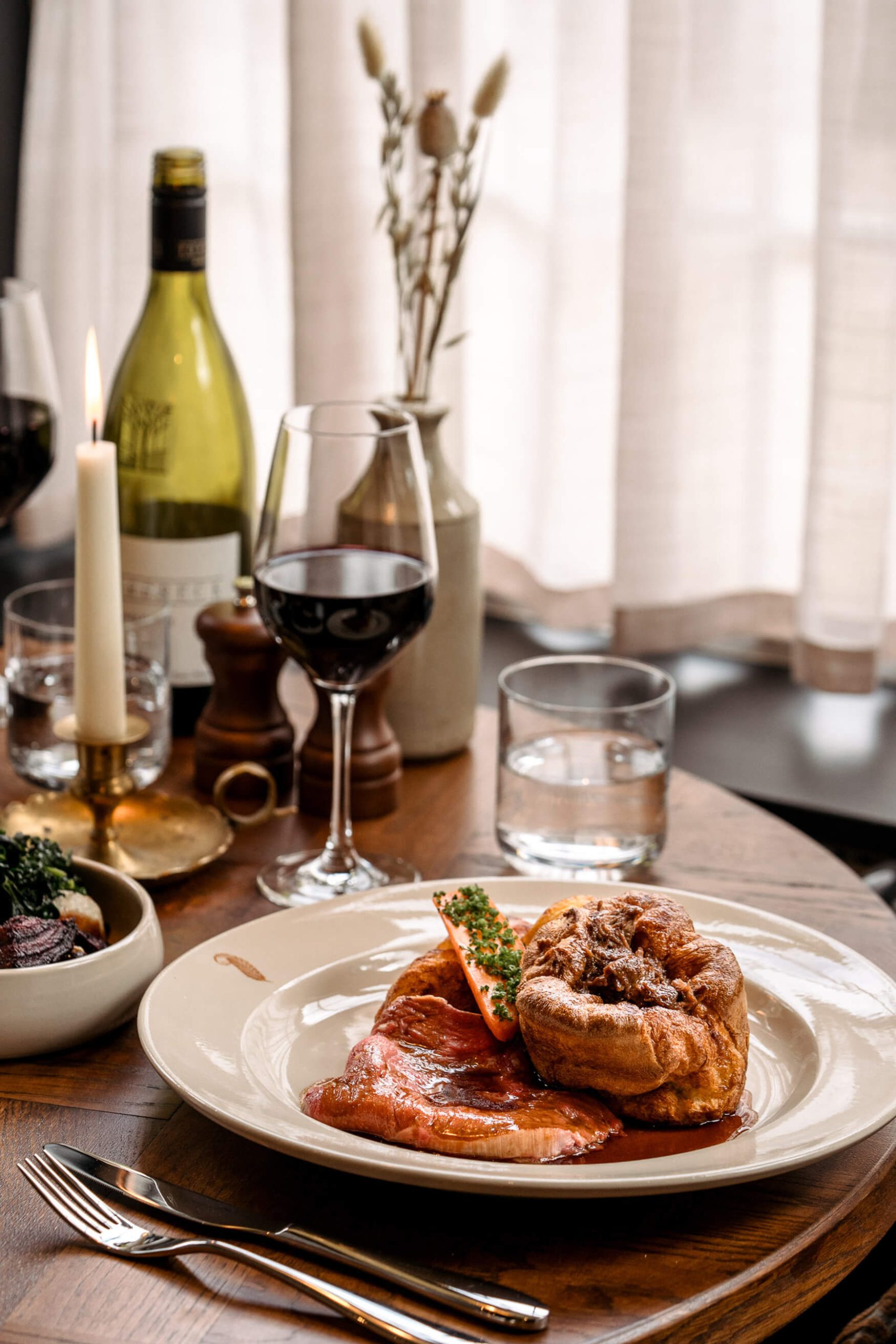 Sunday roast on a white plate sitting on a wooden table amongst all the trimmings and a bottle of red wine